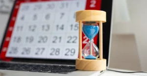 Hourglass with blue sand in front of a computer screen displaying a calendar—representing the importance of timing, scheduling, and commitment in the recovery process at the rehab center.
