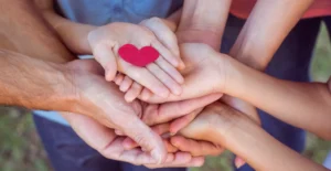 Multiple hands gently layered together holding a small red heart—symbolizing compassion, family support, and the community of care provided by the rehab center.