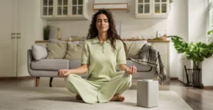 A young woman with curly hair sits cross-legged on the floor in a peaceful living room, meditating with her eyes closed. She wears a light green outfit, and the room features a cozy couch, warm lighting, and lush green plants, creating a serene and calming atmosphere.